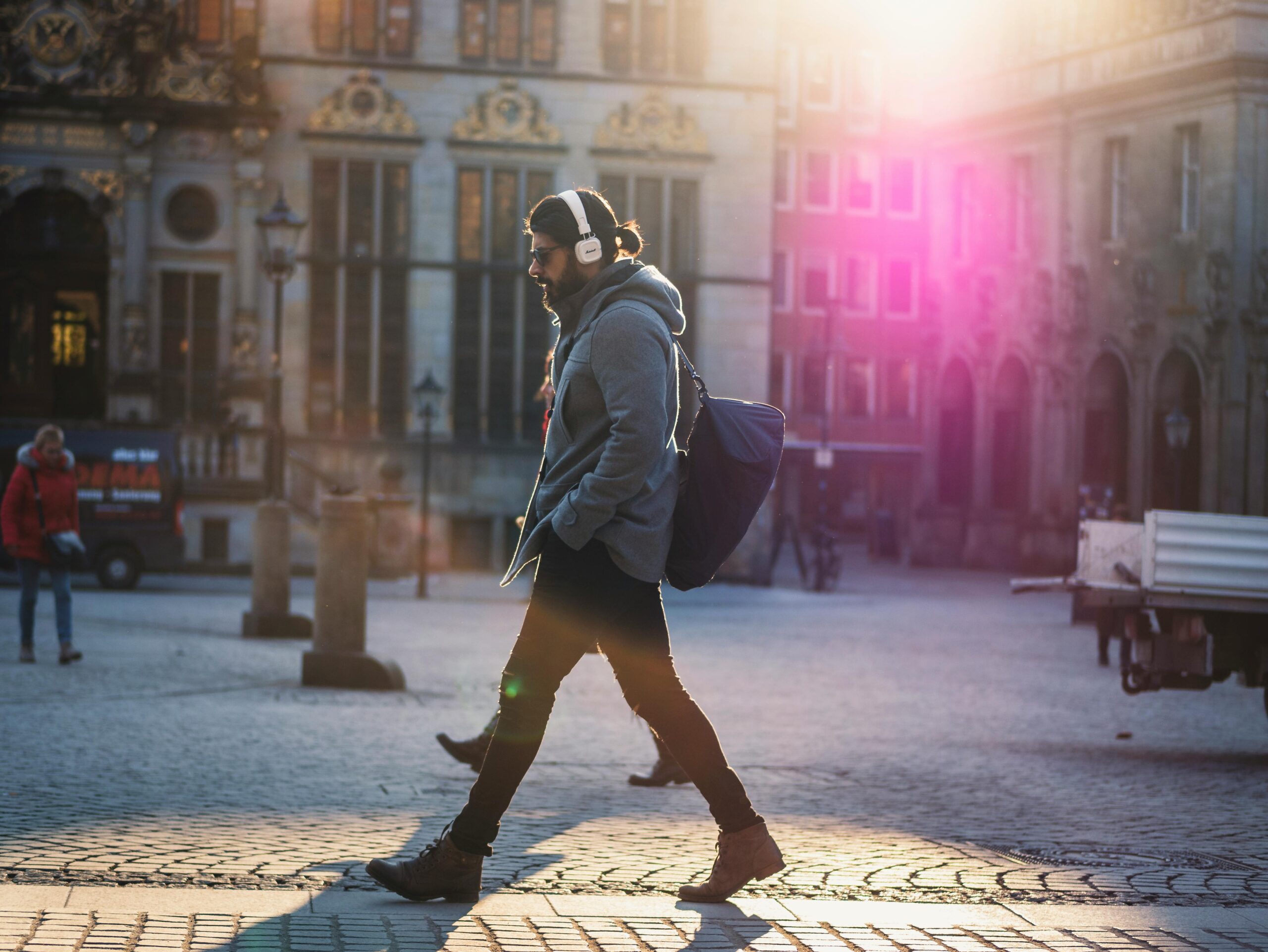 Man with headphones walks down a sunlit urban street, casting a shadow on the cobblestones.